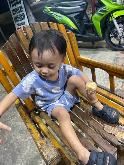 A young child sits on a rustic wooden bench, holding a piece of peeled banana. The child is wearing a blue short-sleeved shirt and shorts with a sheep pattern. Black sandals are on the child's feet. The bench is outdoors, with a concrete floor visible. In the background, a bright green motorcycle is parked. A sign with Indonesian text is also visible behind the child. The scene appears to be located in the city of Ta', Indonesia, possibly an outdoor seating area or near a shop.