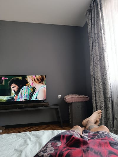 The image depicts a cozy indoor scene, likely a bedroom, with a person lying on a bed. The person's legs and tattooed thighs are visible in the foreground, covered by patterned shorts. In the midground, a television is on, displaying a scene with two people. The room has gray walls and is illuminated by natural light filtering through a large window covered by a patterned curtain. A laundry basket with folded towels sits near the wall, and a power outlet is visible. The overall impression is one of relaxation and leisure, possibly in the evening or during a lazy afternoon, given the soft lighting and the act of watching television from bed. The location is identified as Sandanski, Bulgaria.