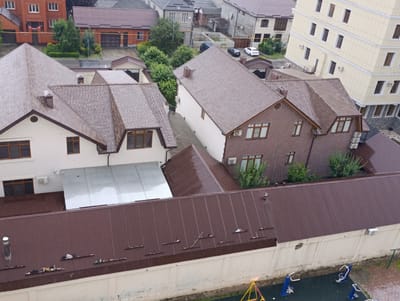 The image shows an aerial view of a residential area with multiple buildings and rooftops. In the foreground, a long, dark brown corrugated metal roof spans across the bottom of the image, topped with a white concrete wall. Several shelves are attached to the wall, holding various objects including what appear to be tools and containers. To the left of the wall, a small playground with yellow and blue structures is visible.

Behind the foreground roof, several houses with dark brown, tiled roofs are prominent. One house in the center is constructed with red brick and white trim, featuring multiple windows. Another larger house to the left has a white facade and a similarly dark tiled roof. Further in the background, other buildings can be seen, including a tall, light-colored building with numerous windows, and red brick houses with dark roofs. Trees and greenery are scattered throughout the scene, indicating a suburban or semi-urban environment. The ground appears wet, suggesting recent rainfall. The overall impression is of a quiet, residential neighborhood. The location is identified as Grozny, Russia.