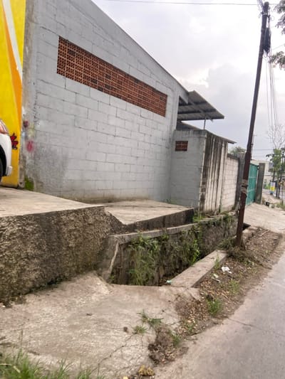 The image shows a concrete wall with a small window and a concrete platform in front of it. The wall is made of cinder blocks and is painted white. The window is made of red brick and is covered with a metal grate. The platform is cracked and uneven, and there is a small ditch in front of it. The ditch is filled with dirt and weeds. The wall is situated in Kecamatan Margaasih, Indonesia, at coordinates (latitude: -6.964, longitude: 107.54). A pole with a black wire on it is in the lower right corner of the image. The street is paved with concrete, with a small amount of debris. There are green plants growing in the ground around the edge of the street.