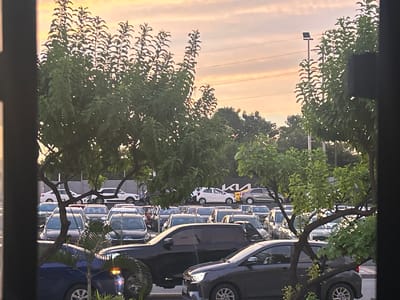 The image shows a parking lot scene in Guayaquil, Ecuador, viewed through a window. A KIA sign is visible. The parking lot is filled with cars of varying makes and models. Trees partially obstruct the view of the vehicles and the sky. The sky suggests it's near sunset or sunrise, with a mix of clouds and a warm color palette. A street light is visible above the trees. The framing suggests the photograph was taken from an interior space. The scene is static, with no visible actions or interactions.
