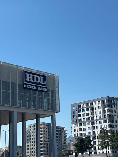 The image depicts a low-angle view of urban architecture in Bjeliši, Montenegro. A prominent building displays the signage "HDL RETAIL PARK" vertically oriented. Behind this structure are several taller apartment buildings with a repeating pattern of windows, indicating a residential or commercial use. The sky is clear and blue. Trees are partially visible within the gaps of the buildings. The overall impression is one of modern urban development in Bjeliši.