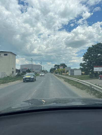 The image captures a street view in Voyvodinovo, Bulgaria, oriented vertically. The scene is dominated by a road with cars, buildings, and a sky filled with dramatic clouds.
In the foreground, a dark-colored car with a yellow sticker on its windshield is driving on the right side of the road. Another white car is visible further ahead on the road. The road is bordered by a grass verge and a white metal guardrail on the left. On the right side of the road, there are buildings, including a modern building with "ARMA" written on it, and a smaller residential building. Signs are visible along the road, one of which reads "Калековец Kalekovets" with a "50" speed limit symbol below it. Trees and utility poles are also present in the scene.
The sky is filled with large, fluffy white clouds against a bright blue sky, suggesting a sunny day with some cloud cover. The time of day appears to be daytime, likely late morning or early afternoon, based on the lighting. The overall atmosphere seems to be one of travel or transit, with the perspective suggesting the photo was taken from inside a vehicle.