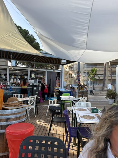 The image shows an outdoor cafe area with white fabric canopies stretched overhead, providing shade. The cafe is located on a wooden deck. In the background, buildings suggest an urban environment, with palm trees and other vegetation. A bar area is visible with shelves stocked with bottles and two women working behind it. Several tables and chairs are arranged on the deck, some of which are occupied. The scene captures a relaxed atmosphere of a cafe in Cambrils, Spain.