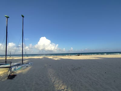 The image shows a beach in Playa del Carmen, Mexico, under a clear blue sky with a large, white cumulus cloud. Two Hobie catamarans with their masts up stand on the white sandy beach. The ocean is a turquoise color with small waves breaking near the shore. Several people are visible in the distance on the beach near the water. The sand shows tire tracks and footprints.