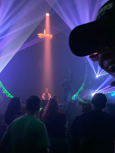The image depicts an indoor event with a DJ booth at the center, illuminated by dramatic lighting. A DJ, wearing a jacket, is seen behind the booth, likely performing. Above the DJ, a red sign with the word "SPARTA" is prominently displayed, lit by a spotlight. The venue appears to be a club or concert hall, with a crowd of people in the foreground, silhouetted against the stage lighting. The atmosphere is dark, with vibrant purple and blue lights creating beams that fan out across the space. The overall scene suggests a lively nightlife setting in South Jakarta, Indonesia.