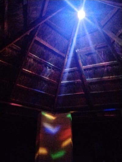 The image shows the interior of a thatched roof structure, possibly a hut or an open-air pavilion, in Mangu Town, Nigeria. A bright light source, emitting blue rays, is visible at the apex of the conical roof, casting dramatic shadows and illuminating the wooden beams and thatch. The overall ambiance suggests a nighttime setting, possibly an event or gathering, as indicated by the colored lights projected onto a central, square-ish pillar. These lights, in shades of yellow, blue, red, and green, create a festive and dynamic atmosphere. The structure appears rustic, with rough-hewn wooden supports and a natural thatch roof, typical of traditional architecture in some rural areas. The scene evokes a sense of community and celebration, with the colorful lights suggesting music, dancing, or some form of entertainment. The dark background suggests it is nighttime, and the light from the central fixture is the primary illumination, supplemented by the colored projections. There is no visible text in the image.