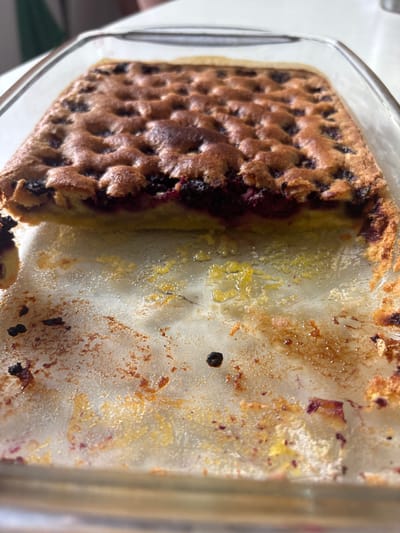 A close-up, high-angle shot shows a berry crumble in a clear glass baking dish. The crumble is golden brown with visible berries peeking through the top. The baking dish has remnants of the crumble, including some burnt sugar and berry stains, indicating it has been recently used. The background is blurred, but appears to be a domestic setting. No specific location in Падлабенскі сельскі Савет, Belarus is discernible from the image.