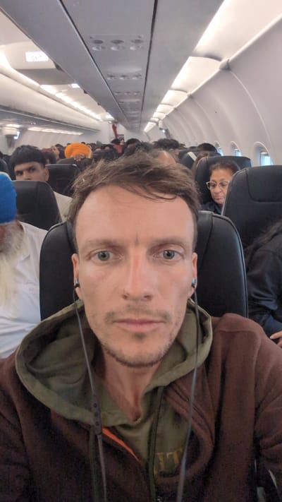 A sideways selfie captured inside a commercial airplane cabin shows the primary subject, a man with light brown hair, stubble, and light green eyes, in the foreground. He looks directly at the camera with a neutral expression, wearing a dark brown hoodie over an olive green hooded garment, where faint letters resembling "LL" or "ALL" are visible. He has earphones in his ears.

The background reveals the interior of the airplane, with rows of dark-colored seats occupied by numerous passengers. Directly behind the main subject, a person with a full white beard, wearing a white shirt and a blue cap or turban, is partially visible. Beside them, another passenger with dark hair and eyes looks towards the camera. Further down the aisle, a passenger wearing glasses is visible. The cabin is brightly illuminated by overhead lights, and white overhead compartments line the ceiling. The setting indicates a full flight, with passengers seated. The activity appears to be a casual moment captured during a flight, possibly while taxiing, cruising, or during boarding/disembarking. Given the location context of Sepang, Malaysia, the flight is likely associated with Kuala Lumpur International Airport. No explicit time of day or weather conditions are discernible from the interior view.