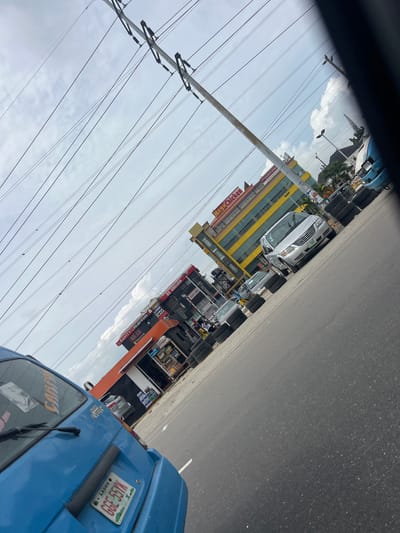 An eye-level shot captures a bright, cloudy day in Way, Nigeria, looking down a road lined with businesses and parked vehicles. Power lines crisscross the sky, supported by poles that stand near the road. On the right side of the road, a blue minivan is parked, facing away from the camera. Its license plate reads "LAGOS GGE 55YK." Further down the road, a silver minivan is parked on the left. Several businesses with prominent signage are visible, including "Mega Chicken" and "DC English." Stacks of tires are piled high along the roadside, suggesting a tire vendor or a repair shop. The road itself is asphalt, with white lane markings visible. The environment appears to be an urban commercial area, with a mix of businesses, vehicles, and infrastructure.