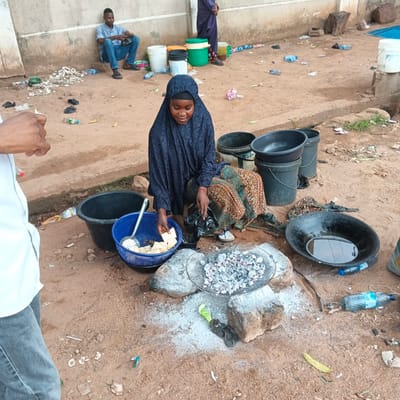 In Shaba, Nigeria, a young girl wearing a dark blue hijab and a patterned skirt kneels on the dirt ground, tending to a cooking setup. Before her, a round metal sieve filled with hot charcoal sits on large rocks, surrounded by a white powdery substance, likely ash or salt. To her left, a blue plastic basket contains white cubes, possibly food items, and a metal strainer. Next to the basket, a large black plastic bucket and a black, shallow pan with water are visible. A person's lower body, wearing gray pants and a white shirt, is partially visible on the left edge of the frame, suggesting they are observing or interacting with the girl. In the background, a man in a light blue shirt and jeans sits leaning against a concrete wall. Various plastic bottles, discarded items, and larger containers are scattered across the sandy terrain.