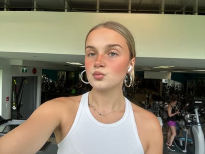 The image shows a woman with blonde hair, blue eyes, a nose piercing, and silver hoop earrings, wearing a white tank top and a silver necklace. She appears to be in a gym setting, as gym equipment is visible in the background, including exercise bikes and rowing machines. Another person is using the exercise bikes in the background. The image appears to be taken at a gym in Whistler, Canada.