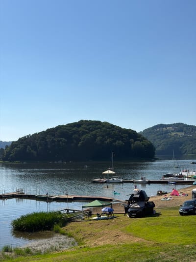 The image depicts a sunny day at a lakeside location, likely Gródek nad Dunajcem, Poland. The scene features a calm body of water, bordered by lush green hills covered in dense trees on the left. On the right, a grassy embankment slopes down towards the water, where a wooden dock or pier is situated.

Several boats are moored along the dock, including sailboats and smaller watercraft. Some of these boats appear to be occupied or have people on board. On the grassy area next to the dock, various objects and activities are visible. These include parked cars, picnic blankets, beach umbrellas, and camping gear, suggesting a recreational day. People can be seen relaxing, walking, and possibly preparing for water activities. One person is visible in the water, appearing to swim or play.

The clear blue sky indicates fair weather, and the bright sunlight suggests it is midday or early afternoon. The overall atmosphere appears relaxed and leisurely, characteristic of a summer outing at a scenic lake. There is no visible text in the image.