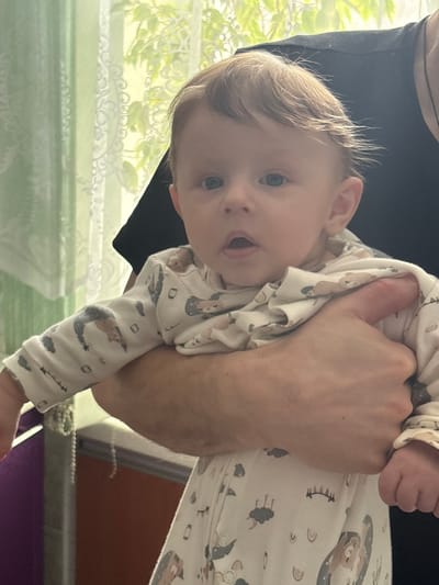 In the city of Furmanov, Russia, an indoor scene displays an adult holding an infant. The infant, positioned centrally, exhibits fair skin, light brown hair, and blue eyes, with the mouth slightly open. The infant wears a white long-sleeved onesie adorned with light-colored patterns, including bears, rainbows, and clouds. A fair-skinned adult's right arm supports the infant's midsection, and a dark garment is visible behind the infant's head. The background features a window covered by light green, patterned sheer curtains, through which green foliage is visible. To the lower left, a segment of a purple object, a light-colored rail, and a vertical string of white beads are present. A reddish-brown horizontal surface occupies the lower background.
