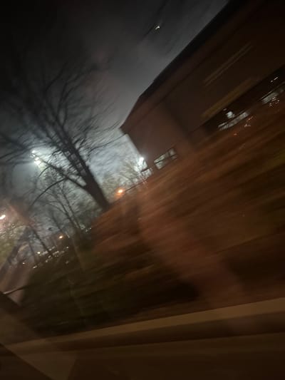 An abstract view appears to be from inside a building, looking out through a window. Silhouetted trees with bare branches reach toward a dimly lit sky. A building with visible windows is partially visible. The image has a blurred, distorted quality, possibly due to motion or focus issues. The overall scene evokes a sense of night, winter, and an indoor-outdoor contrast.