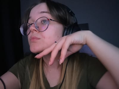 A young adult female is depicted in a close-up, dimly lit indoor setting in Pushchino, Russia. She has light skin, brown hair with visible blonde highlights, and wears round, black-framed glasses with a red accent on the right temple, featuring a small reflection of blue and purple light. A silver stud nose piercing is visible on her right nostril. Black over-ear headphones are worn on her head, with the right earcup partially visible. She wears a dark olive green short-sleeved top. Her left hand is positioned with fingers slightly bent, resting against her lower right face and jawline. This hand displays glittery nail polish and a silver-toned ring with a central dark blue or purple stone surrounded by smaller clear stones on the ring finger. A black elastic band is visible on her left wrist. The background is uniformly dark and out of focus, suggesting low ambient light.