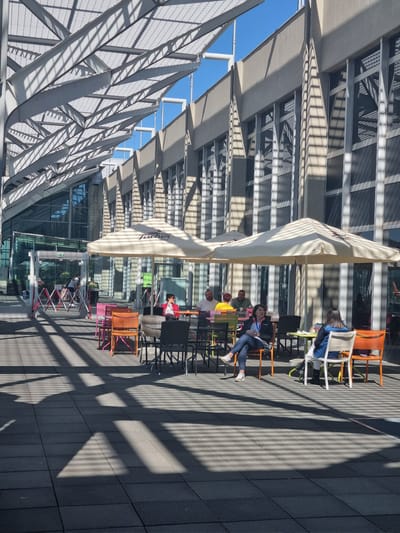 The image depicts an outdoor cafe in Saint-Louis, France. The cafe is situated under a modern architectural structure featuring a geometric metal framework casting linear shadows. Several people are seated at tables, engaged in conversation or relaxation. The cafe furniture includes colorful chairs. An outdoor umbrella with a branded logo provides shade. The ground is paved with dark gray rectangular tiles. The architecture suggests a contemporary urban environment.