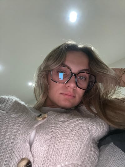A woman wearing oversized square glasses looks at the camera. She has light brown hair that is parted to one side. She's wearing a fluffy cream-colored cardigan with large buttons. The background is a plain wall with several bright light reflections. A small portion of a dark gray pillow is visible behind her head.