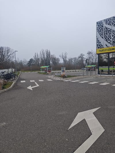 This image captures an outdoor commercial parking area in Hœnheim, France, under an overcast and hazy sky, suggesting low visibility. The foreground features dark grey asphalt pavement with a prominent white L-shaped directional arrow painted on its surface, indicating a left and forward turn. To the immediate right of this arrow, a white dashed-line crosswalk is visible, leading to a textured pedestrian path. On the far left, a grassy median, bordered by large light-colored rocks and a low concrete or stone barrier, accommodates several black bicycles secured together.
In the midground, the asphalt extends into a broader expanse, likely a parking lot. A tall, dark street light post is positioned on the left. Two identical green-roofed structures, appearing to be self-service bays, are situated in the background; one is towards the center-left, and the other towards the center-right. Between these structures, a freestanding sign displays an image of a human figure. A dense line of bare trees forms the distant background, with their branches devoid of foliage.
On the far right, a large building with a distinctive grey and black geometric patterned facade is partially visible. Attached to this building is a bright yellow sign displaying the text "FRUITS ET LÉGUMES" in black capital letters. Multiple vertical metal bollards are installed in front of the building, possibly marking an entrance or pedestrian zone. A dark-colored vehicle is partially discernible in the background, positioned near the rightmost green-roofed self-service bay. No individuals are present, and no discernible actions or interactions are occurring within the scene.