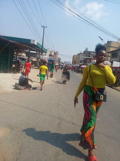 The image depicts a bustling daytime street scene in Nkwelle Ezunaka, Nigeria. The paved asphalt road shows signs of wear, with visible cracks and variations in texture. Strong overhead sunlight casts distinct shadows across the pavement.
In the foreground, slightly to the right of center, a female subject walks towards the viewer. She wears a long-sleeved, sheer yellow top, a long skirt with a vibrant multi-colored pattern (green, red, orange, black), and red open-toed sandals. A small black shoulder bag with a gold horseshoe-shaped clasp hangs from her left shoulder. She holds a dark-colored smartphone in her right hand, while her left hand is raised towards her neck area. Her dark, curly hair is styled upwards.
In the midground, to the left, a barefoot man sits directly on the pavement, dressed in a light blue short-sleeved collared shirt and dark trousers. A small dark bag or bundle rests beside him, and a small blue bowl is on the ground in front. Further down the street, multiple pedestrians are visible, moving away from the viewer. These include an individual in a bright red dress, and another in a yellow t-shirt and green shorts carrying a white container on their head. A dark-colored wheelbarrow is also present, appearing to be pushed or pulled.
The background on the left features a low-lying structure with a green and white striped canopy, possibly part of a gas station or a large market stall. Numerous roadside stalls or small shops line the left side of the street, many with corrugated iron roofs and displaying merchandise. On the right side of the background, multi-story buildings with various architectural details are visible, some featuring satellite dishes. A utility pole supporting multiple overhead power lines stands prominently in the mid-left. The sky above is clear and blue with minimal cloud cover.