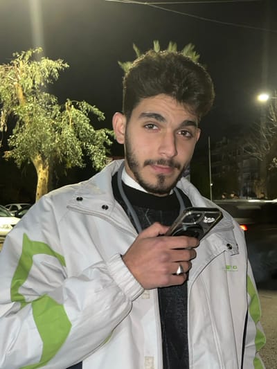 A young man with dark hair and a beard poses outdoors at night. He holds a smartphone to his ear with his right hand, where he wears a silver ring. He is dressed in a white jacket with bright green accents, layered over a dark sweater and a collared shirt. Behind him are dimly lit trees and a building in the background. The scene is lit with soft, ambient lighting.