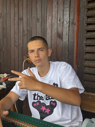 The image shows a young man with a buzz cut, wearing a white t-shirt with a graphic design of a bear and stars, making a peace sign with his right hand. He is sitting at a table covered with a green tablecloth featuring an ethnic pattern. In the background, there is a wooden wall with visible vertical planks, an electrical outlet, and a louvred shutter on the right. The scene appears to be outdoors or in a rustic, covered area, possibly a porch or an outdoor seating area of a building in Vladislavtsi, Bulgaria.