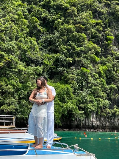 In Ao Nang, Thailand, a couple stands embracing on the deck of a boat. The woman, wearing sunglasses and a floral top with a light blue and white patterned skirt, is positioned in the foreground, her arms wrapped around herself. Behind her, a man in a white outfit stands with his arms around her waist. The boat, with its blue, white, and yellow accents, rests on turquoise water. In the background, a dense, lush green cliffside rises, covered in vegetation and trees. Several other boats are visible, and people can be seen swimming and enjoying the water.