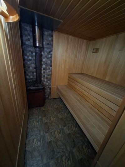 The image shows the interior of a sauna. The walls and ceiling are paneled with light-colored wood. There are two tiers of wooden benches in the right side of the image. On the left side of the image, a metal stovepipe extends from a dark red metal stove, which is set against a wall of small, square mosaic tiles. The floor is covered with dark, textured tiles. There are no people visible in the image. The location is described as being in the city of Kara-Balta, Kyrgyzstan, which is a detail that cannot be verified from the visual information provided in the image.