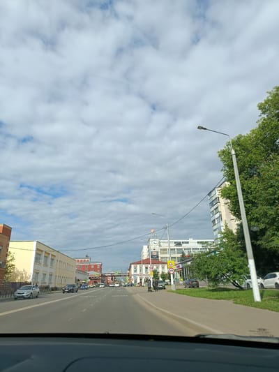 The image shows a street scene in Orekhovo-Zuyevo, Russia, on a cloudy day. A multi-lane road is visible, with cars driving in both directions. On the left side of the road, there is a yellow, two-story building with many windows, and further down, a red brick building. On the right side, a modern apartment building with balconies is seen, alongside trees and a sidewalk. A tall streetlamp stands prominently on the right. Several traffic signs are visible, including speed limit signs indicating 40 and 20. People are walking on the sidewalk, and a person is seen pushing a stroller. In the distance, more buildings and infrastructure, possibly a bridge, can be observed. The sky is filled with a mix of white and grey clouds.