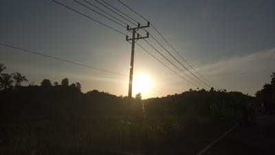 This image captures a sunset scene in Seuneubok, Indonesia. A prominent utility pole with multiple power lines stretches across the frame, silhouetted against the bright, setting sun. The sun, a brilliant orb, is partially obscured by the utility pole and a cluster of trees on a gently sloping hill in the background. The foreground is dark and features tall grasses. The sky is a gradient of blue and pale orange, with some wispy clouds visible. The overall impression is a rural or suburban landscape at dusk, with the power lines indicating the presence of human infrastructure in a natural setting. There are no discernible people or specific actions depicted, but the scene suggests the end of the day.