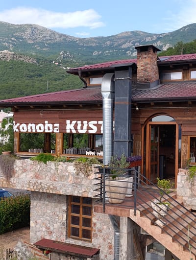 The image shows a restaurant named "Konoba Kust" located in Kamenovo, Montenegro. The restaurant is built with wood and stone, and it is situated on a hillside with mountains in the background. The restaurant has a large outdoor seating area with tables and chairs, and a terrace with potted plants. There is also a large chimney and a metal pipe on the exterior of the building. The sky is blue with some clouds.
