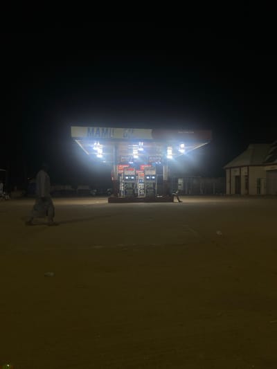 The photo shows a MAMI petrol station in Unguwar Fatika, Zaria, Nigeria, taken at night. The station is illuminated by bright lights from the canopy. Petrol pumps are visible under the canopy. A figure in light-colored clothing stands in front of the station. The environment is dark, except for the immediate vicinity of the station. The building has the words “Kafan Doka Zaria” on the canopy.