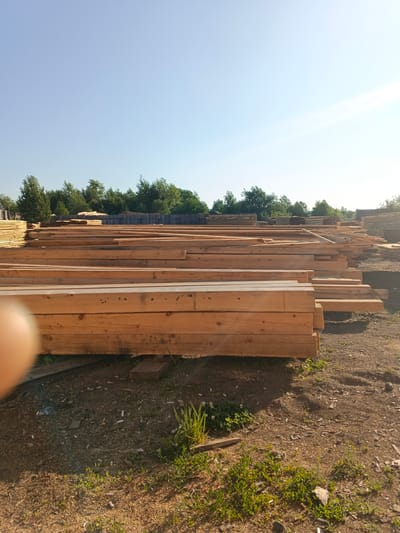 The image depicts an outdoor lumberyard under a clear blue sky with the sun casting a warm glow. In the foreground, large stacks of raw, unfinished wooden planks are piled high. These planks are light brown and appear to be of uniform size, suggesting they are for construction or manufacturing. Behind the main stacks of lumber, more piles of wood can be seen, along with a wooden fence and a dense line of green trees forming a natural backdrop. The ground is covered with dirt, wood chips, and scattered debris, including cigarette butts and small stones. The overall scene suggests an industrial or storage area for lumber, likely in a rural or semi-rural setting. The location is specified as the city of Strelka, Russia. There are no people visible in the image.