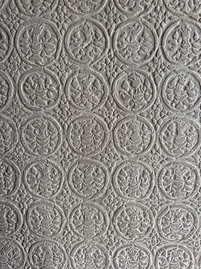 The image shows a close-up view of a textured surface with a repeating pattern of embossed circular motifs. Each motif appears to contain a stylized floral or figural element within a circular frame, with smaller floral patterns filling the spaces between the larger circles. The overall impression is of an ornamental design on a material such as stone, plaster, or possibly embossed paper or fabric. The lighting creates shadows that highlight the relief of the pattern.
This pattern is characteristic of carvings found on the walls of ancient temples in Siem Reap, Cambodia, particularly within the Angkor Archaeological Park. Such intricate bas-reliefs are a hallmark of Khmer architecture and artistry, often depicting religious scenes, mythological figures, or decorative motifs. For example, this style of carving is frequently seen on the walls of Angkor Wat, Bayon, and Ta Prohm temples, where they contribute to the rich historical and cultural narrative of the region.