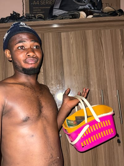 The image depicts an adult male standing indoors, identified as being in Muchiya, Nigeria. The male is bare-chested, exhibiting dark skin and visible dark body hair on his chest and abdomen. He wears a dark blue knit beanie with a lighter pattern. He has a short beard and full lips, slightly parted. His gaze is directed upwards and to the viewer's right; his left eye shows a distinct red reflection, consistent with a camera flash effect.
In his left hand, he holds a vibrant multi-colored plastic basket. The basket's main body is hot pink with vertical slatted sides, featuring a yellow/orange rim and interior, and two white plastic handles. Visible inside the basket are what appear to be several green and white currency notes and a dark, rectangular object.
The background consists of a wooden wardrobe or cabinet with vertical wood grain patterns. On the right side of the wardrobe, two vertical metallic handle bars are present, and a set of keys hangs from a small fixture. Resting on top of the wardrobe are various items including multiple pairs of sandals or slippers, dark fabric or clothing, and a cardboard box partially labeled "A4 SERIES" and "NATURAL COLOUR ENHANCEMENT," with additional text "DTS VIRTUAL:X" also visible. The scene is illuminated by artificial light.