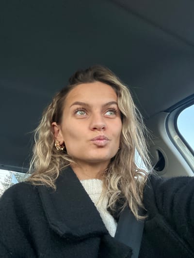 A woman with blonde, wavy hair poses in a car. She wears a black coat over a white sweater and gold earrings. She is turned slightly to her left with her lips pursed in a kissing face. The car's interior is visible, including the ceiling and a seatbelt across her shoulder. Natural light illuminates her face and highlights her green eyes.
