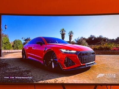 The image displays a red Audi RS7 parked on a textured surface, possibly asphalt or gravel, with a backdrop of lush greenery and palm trees under a clear blue sky. The car is prominently featured in the center, showcasing its sleek design and aggressive styling. In the lower left corner of the screen, there are interface elements indicating game controls: a "Save" button with a PlayStation controller icon, an "Exit" button with a circle icon, and a "Change Watermark Position" button with a keyboard icon. In the lower right corner, the "FORZA HORIZON 5" logo is visible. The scene appears to be a digital rendering from a video game, specifically Forza Horizon 5, which is set in Mexico, not Sevlievo, Bulgaria. There are no people or actions depicted.