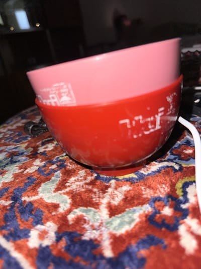 The image shows two plastic bowls, one pink and one red, stacked on top of each other. The pink bowl is on top, and the red bowl is below it. Both bowls have some sort of white lettering or design on them. The bowls are resting on a patterned surface, which appears to be a rug with a mix of red, blue, and white colors. In the background, there is a dark, out-of-focus area that could be furniture or a wall. A white cable is visible extending from the right side of the image, partially obscured by the red bowl. The overall lighting is dim, with some reflections visible on the surfaces of the bowls. There are no people visible in the image, and no identifiable landmarks or specific details that would indicate a location in Gyumri, Armenia.