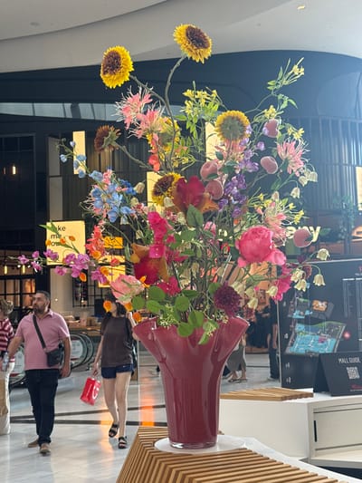 The image captures a vibrant floral arrangement in a large red vase, prominently displayed indoors. The flowers are a mix of colors including yellow, pink, purple, and blue, with some resembling sunflowers, peonies, and delphiniums. Small, peach-colored decorative fruits are interspersed among the blossoms. The background suggests an indoor public space, possibly a shopping mall, with architectural elements like a curved white wall and vertical wooden paneling.
In the background, several people are visible. One woman in a brown top and blue shorts is walking, holding a red shopping bag. Another person, wearing a pink polo shirt and dark pants, is walking towards the right side of the frame. A third person, wearing a striped shirt, is also visible in the lower part of the image. The lighting indicates it is daytime. A sign with "MALL GUIDE" and a map is also visible on the wall, further suggesting a mall setting. The overall impression is one of a well-decorated public space.
