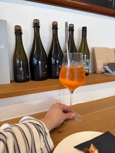 The scene is an indoor setting that appears to be a wine bar or tasting room in Meisenheim, Germany. A hand is holding a wine glass filled with an orange-colored drink, possibly an Aperol spritz or similar cocktail. The glass is positioned over a horizontal display of wine bottles arranged on a shelf against a white wall. Five bottles are visible, with dark green glass and black labels, suggesting sparkling wine or similar. A metal straw is inserted into the glass. Behind the bottles is a wooden shelf. To the right, a white plate with a small food item on top of a black napkin is partially visible. A chalkboard and some papers are also slightly visible on the left side.