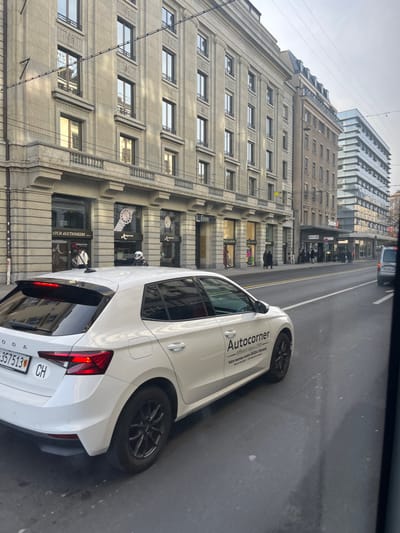 The image captures a daytime street scene in an urban environment, likely in Geneva, Switzerland, viewed from inside a moving or parked vehicle.
In the foreground, a white, modern-looking hatchback car, identified as a Skoda by the logo on its trunk, occupies the right side of the frame. The car has "Autocorner" written in black text on its side, along with smaller, unreadable text below it. The Swiss license plate "CH VD 357513" is clearly visible on the rear. The car features black multi-spoke wheels.
To the left of the street, a tall, classical-style building with many floors and numerous rectangular windows dominates the mid-ground. Its facade appears to be made of light-colored stone or concrete, adorned with architectural details. The ground floor of this building houses several retail storefronts, including one labeled "WATCH AUCTIONEERS" and another partially visible as "Bankor." Display windows showcase various items, including a large watch face. A few indistinct figures, likely pedestrians, can be seen on the sidewalk in front of these shops, one appearing to stand near a storefront or entering it. Another person on a small scooter or motorcycle is visible near the sidewalk.
Further down the street, behind the classical building, a more contemporary building with a distinctive glass and metal facade is visible, reflecting the overcast sky. The road surface is asphalt, appearing slightly damp in places. Overhead power lines or tram wires stretch across the top portion of the frame. The sky is uniformly gray and overcast, suggesting a cloudy day with diffused light. The overall scene depicts typical city life with traffic and commercial activity.