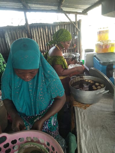 This image depicts an informal food preparation and vending stall in Cotonou, Benin. Two adult women are actively engaged in tasks within the semi-open structure, which features a corrugated metal roof and wall supported by rustic wooden poles.

In the foreground, a woman wearing a teal headscarf with light embellishments and a matching top over a patterned fabric wrap is bent over, focusing on items within a pink plastic basket containing green and white patterned plates or bowls. Her hands are positioned as if handling the contents.

In the background, a second woman, wearing a green patterned headscarf tied at the back and an off-the-shoulder green patterned top, stands behind a counter. She holds a dark object in her right hand above a silver metal colander or bowl. Adjacent to her on the counter is a large, silver cooking pot with a partially open lid, resting on a white tarp draped over a patterned fabric. The pot contains a dark liquid or sauce, along with several lighter, spherical items and darker, elongated items. To the right of the background woman, stacks of plastic bottles containing yellow-orange and pink liquids are visible, alongside a blue and grey portable cooler. Bright light enters the scene from the right, illuminating the area.