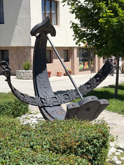 The image shows a sundial sculpture in the city of Bansko, Bulgaria. The sundial is made of dark metal and is shaped like an anchor. It is located in front of a building with a white facade and stone accents. There are green bushes in the foreground and a tree with green leaves to the right of the sculpture. The ground is paved with stone tiles. The sundial has Roman numerals and possibly Cyrillic letters on its surface. The year "2019" is visible at the bottom of the sculpture. There are no people clearly visible in the immediate vicinity of the sundial.