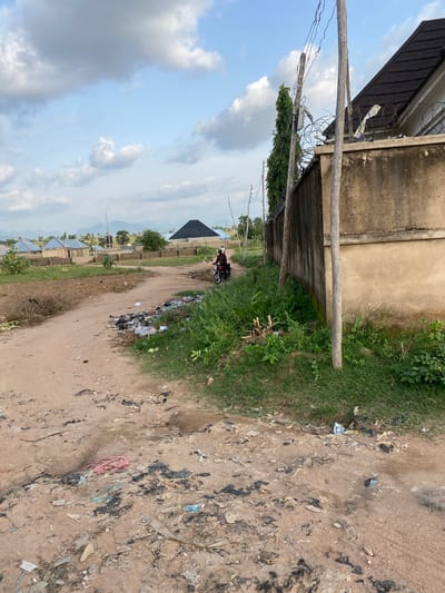 The image depicts a dirt road in Kuson, Nigeria, with scattered debris and garbage along its edges and in the foreground. The road curves towards the left of the frame, with a stone wall and a building on the right side, topped with barbed wire. In the distance, several houses with dark, angular roofs are visible, set against a backdrop of rolling hills under a partly cloudy sky. Two individuals are riding a motorcycle on the road, moving away from the camera. Lush green vegetation grows alongside the road and in front of the wall.