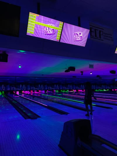 The image depicts the interior of a bowling alley in Waynesboro, United States of America, bathed in neon purple and green lighting. Two large screens hang from the ceiling. The left screen displays a bowling score with names like Oscar, Tai, Fer, Casey, Shani, Romina, and Adi. The right screen shows a cartoonish television with a logo that reads "You Rookies." A banner for "ECP Engineered Construction Products LTD." is visible on the right wall. Several bowling lanes with illuminated pink and green borders stretch into the distance. A person, silhouetted against the light, is standing on one of the lanes, facing away from the camera. In the foreground, a bowling ball return machine is partially visible. The overall scene suggests a vibrant and lively atmosphere typical of a bowling entertainment venue.