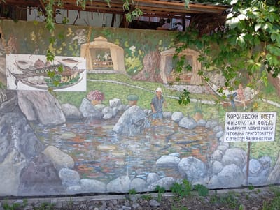 This image depicts a painted mural, likely on a wall. The mural features a scene depicting a pond or river with rocks, fish, and a person wading in the water holding a net. In the background, there are depictions of two small tent-like structures, a couple in a hammock, and foliage. A sign is present on the right side of the mural in Cyrillic, which translates to "Royal Sturgeon and Golden Trout. Choose any fish, and the chefs will cook it to your liking." The upper part of the mural is partially covered with hanging plants and a wooden structure, suggesting the mural is outdoors and possibly part of a restaurant or recreational area. Chinarkent, Uzbekistan, is noted as the location.