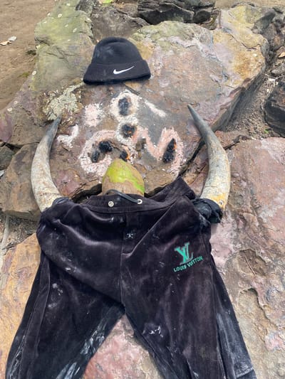 The image depicts an outdoor scene with a collection of objects laid out on rough, rocky terrain. The primary subjects are a pair of animal horns, a coconut, a black beanie with a white Nike logo, and a black velour tracksuit with a green Louis Vuitton logo. The horns are positioned on either side of the coconut, and the tracksuit is draped over them and the coconut. Small piles of what appears to be ash or charcoal are scattered on the ground between the horns.

The setting is rocky and appears to be outdoors, with some dirt and dry leaves visible. The time of day and weather are not explicitly indicated, but the lighting suggests it is daytime. There are no people visible in the image.

Notable details include the prominent branding on the beanie and tracksuit, suggesting these are items of clothing. The arrangement of the objects, particularly the horns and coconut with the ash, may indicate a ritualistic or symbolic placement, although this is speculative.

Visible text includes the "NIKE" logo on the beanie and "LOUIS VUITTON" with the "LV" monogram on the tracksuit. The location context provided is Achara, Nigeria.