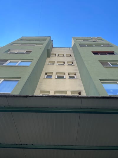 An eye-level, wide shot taken from below shows a light green building against a clear blue sky in Berkovitsa, Bulgaria. The building is viewed from its base, looking up. The building is divided vertically into two sections. The left section is painted a light green, while the right section is painted a cream or beige color. Both sections are multistory, with rows of windows visible. Some windows are open, while others are closed. The building has a diagonal architectural feature where the two colored sections meet. The bottom of the image shows a section of a covered walkway or overhang, with a corrugated white ceiling and green metal supports. The overall impression is a modern, somewhat stark architectural style.