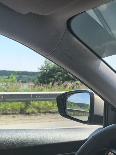 The image shows the interior of a car looking out the passenger window. A side-view mirror reflects a landscape of trees, a body of water, and a blue sky. The word "AIRBAG" is visible on the car's interior trim. The scene suggests a journey through a rural or semi-rural area. The location in Sysert, Russia is not identifiable from the image content.