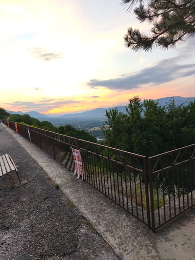 The image captures a scenic view from a high vantage point overlooking a valley, possibly near Bojano, Italy, at sunset. The sky is ablaze with warm hues of orange, yellow, and pink, transitioning into a pale blue at the top, with scattered clouds. In the foreground, a brown metal railing lines a concrete pathway. A wooden bench with white slats is visible on the left, situated on a gravelly surface. Two small, white flags with red cross designs are hanging from the railing. Beyond the railing, lush green trees and vegetation dominate the middle ground, with a town or village nestled in the valley below. In the distance, several layers of mountains recede towards the horizon, bathed in the soft light of the setting sun. A pine branch hangs into the frame from the top right corner.